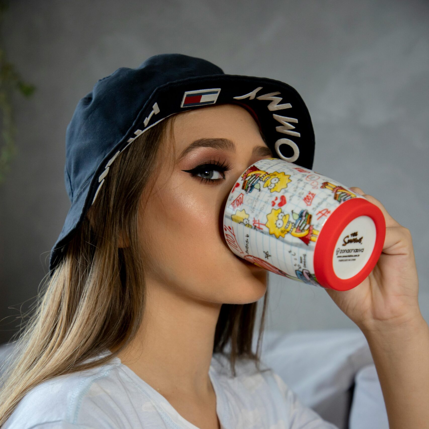 Adult woman sipping from a colorful cartoon mug wearing a trendy bucket hat indoors.
